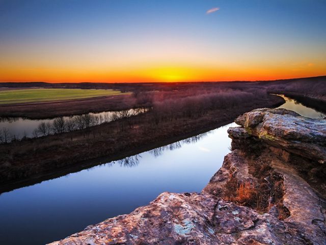 A river running between a rocky bluff and flat grassy fields with a yellow and orange sunset in the distance.