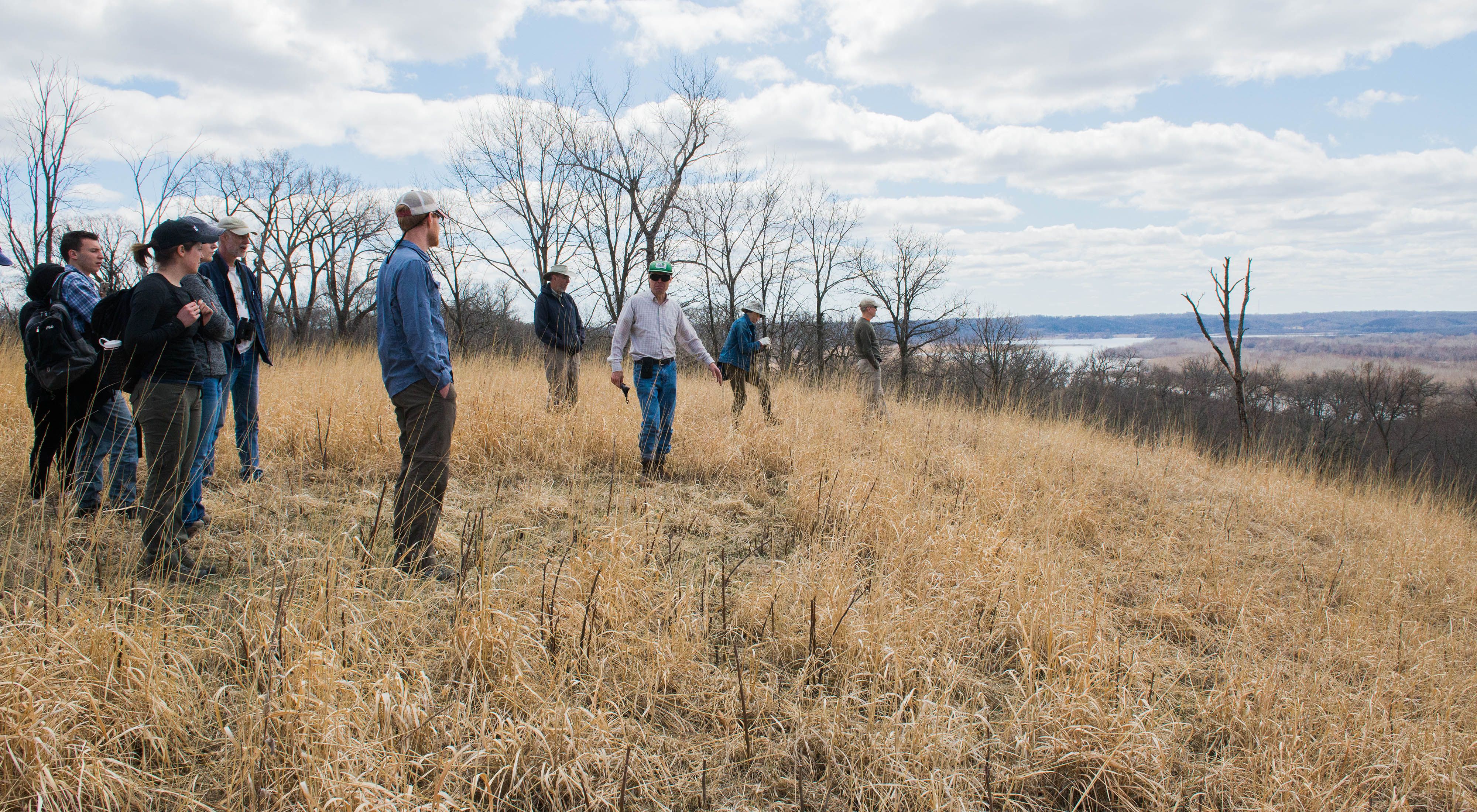 A group of people stand on a blufftop covered in golden grasses and look out toward a river.
