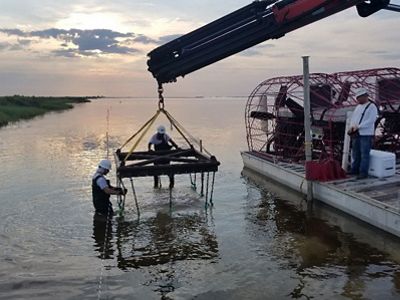 A frame for oyster reef restoration being lowered into the water by a barge-mounted crane.