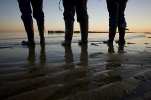 Beach-level view of the boots and lower legs of three people standing at the edge of a body of water.