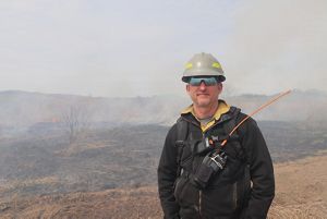 Brian Winters stands in front of a prescribed fire with fire and smoke in an open field.