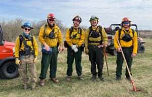 Fire crew members in yellow outfits stand together and smile in an open field.