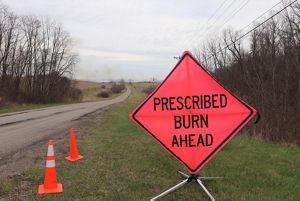 Orange sign reads prescribed burn ahead with smoke in the background.