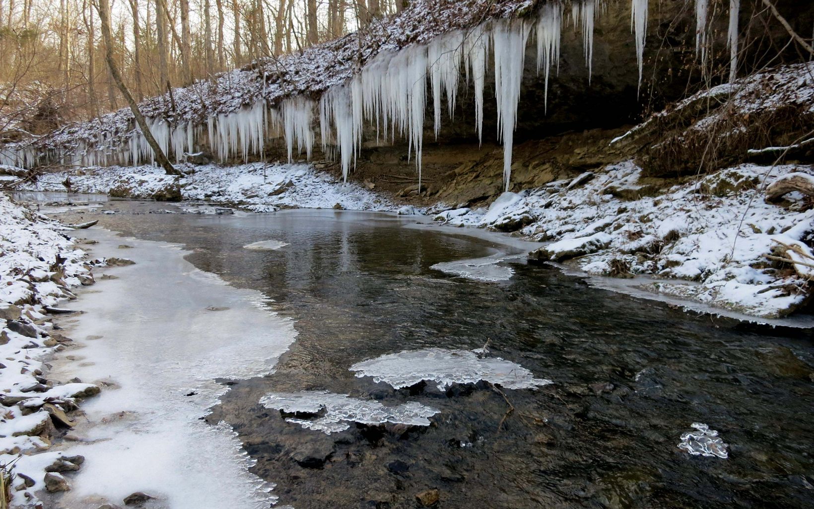 Icicles hang from a rocky outcrop over a partially frozen stream. 