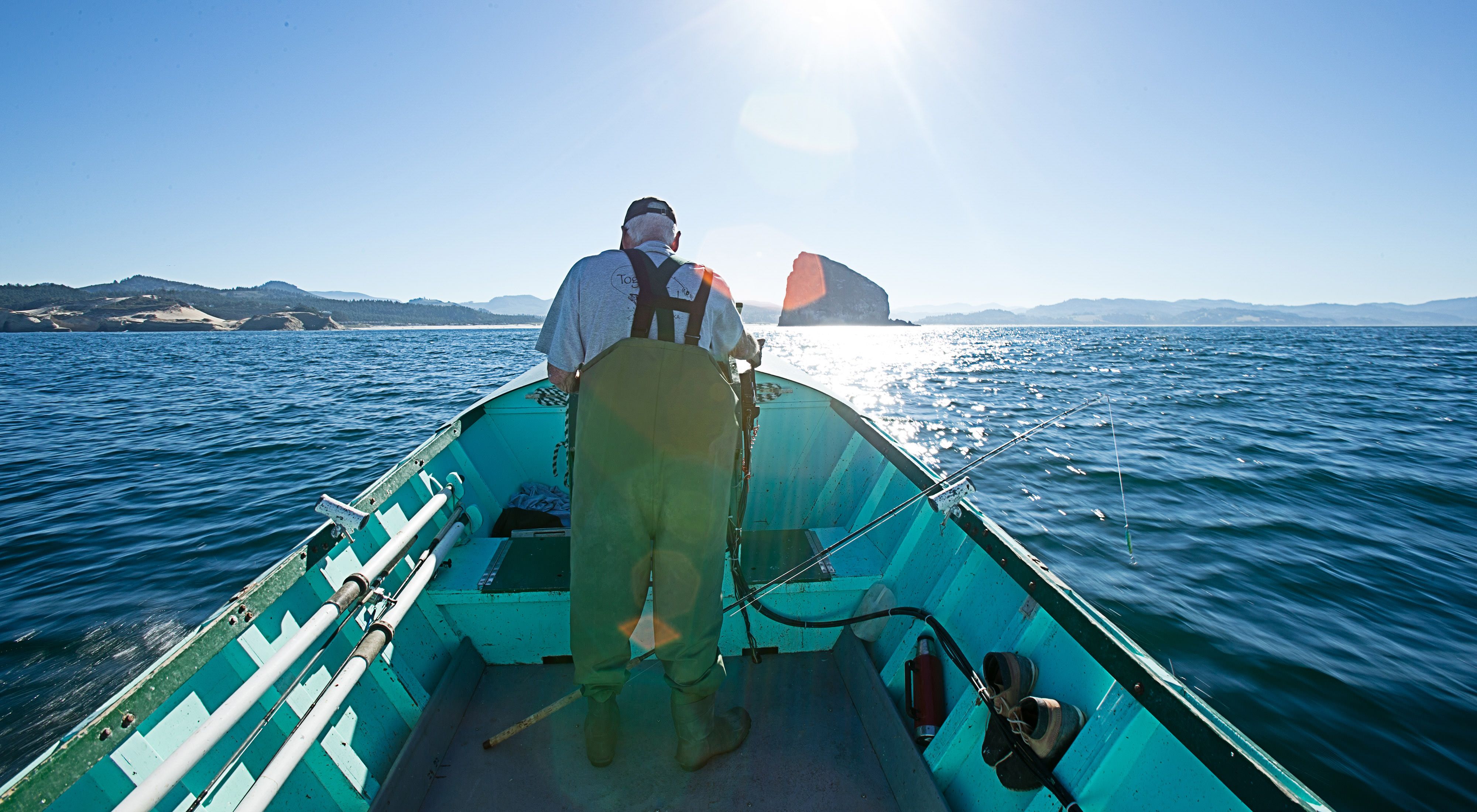 A man wearing waders stands at the bow of a small boat on a wide body of water.