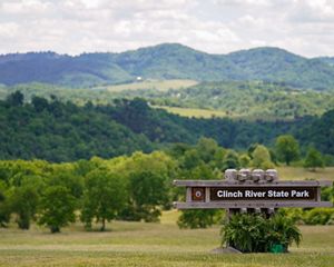 A large sign reading Clinch River State Park welcomes visitors. Rolling tree covered hills rise behind the sign toward a tall mountain ridge that lines the horizon.