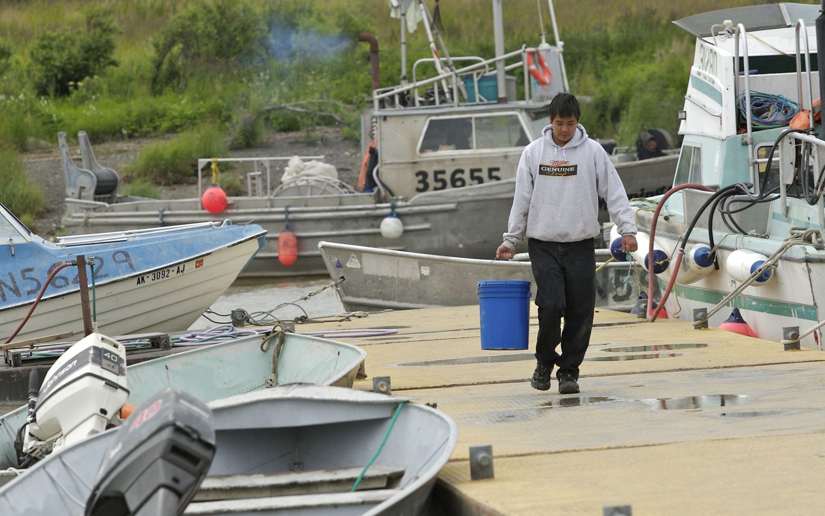 Person carrying a blue pail walking across a fishing dock with boats surrounding the dock.