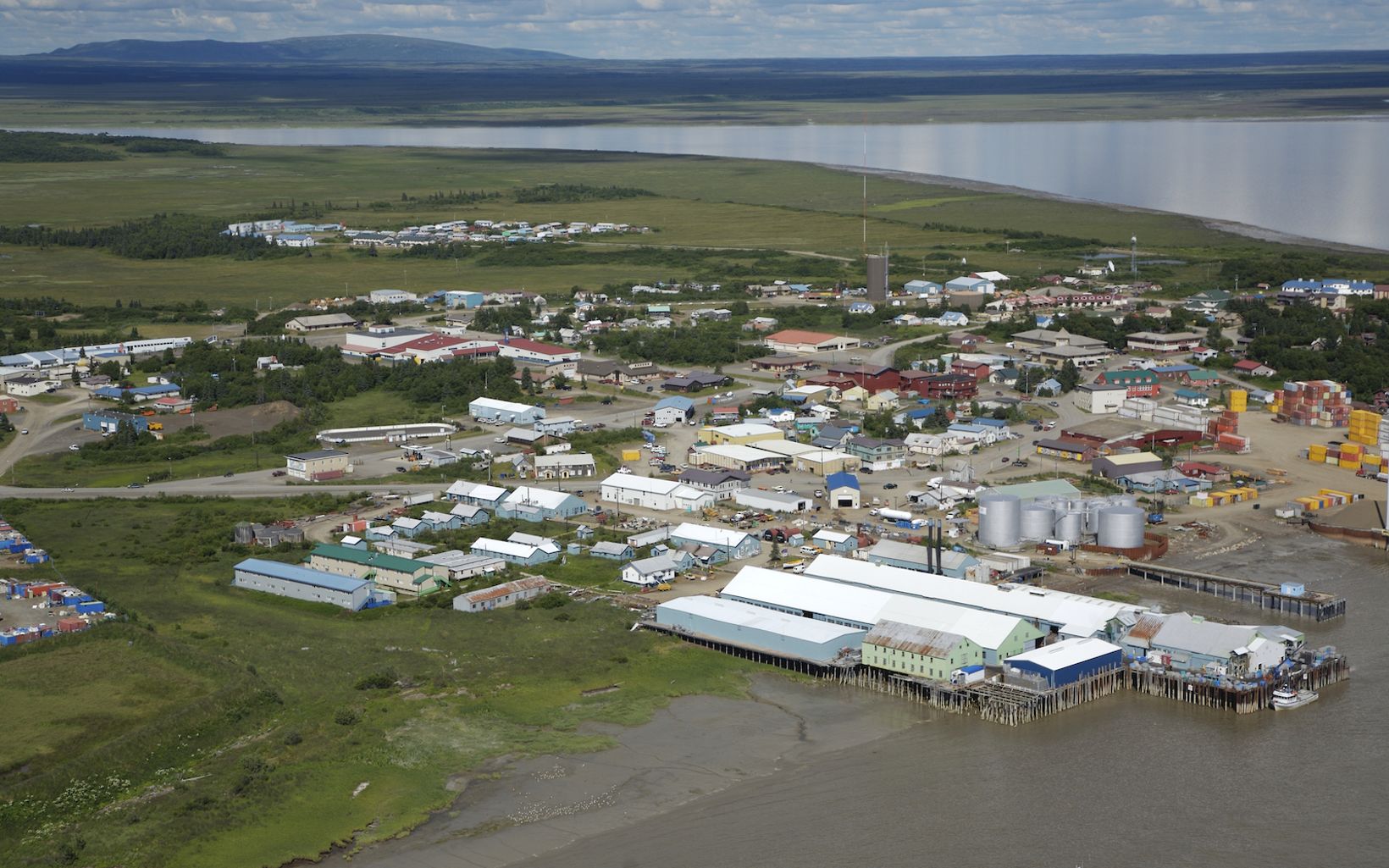 An aerial view of a small fishing town with many industrial buildings near the water, surrounded by the Bristol Bay with shoreline and mountains in the distance.