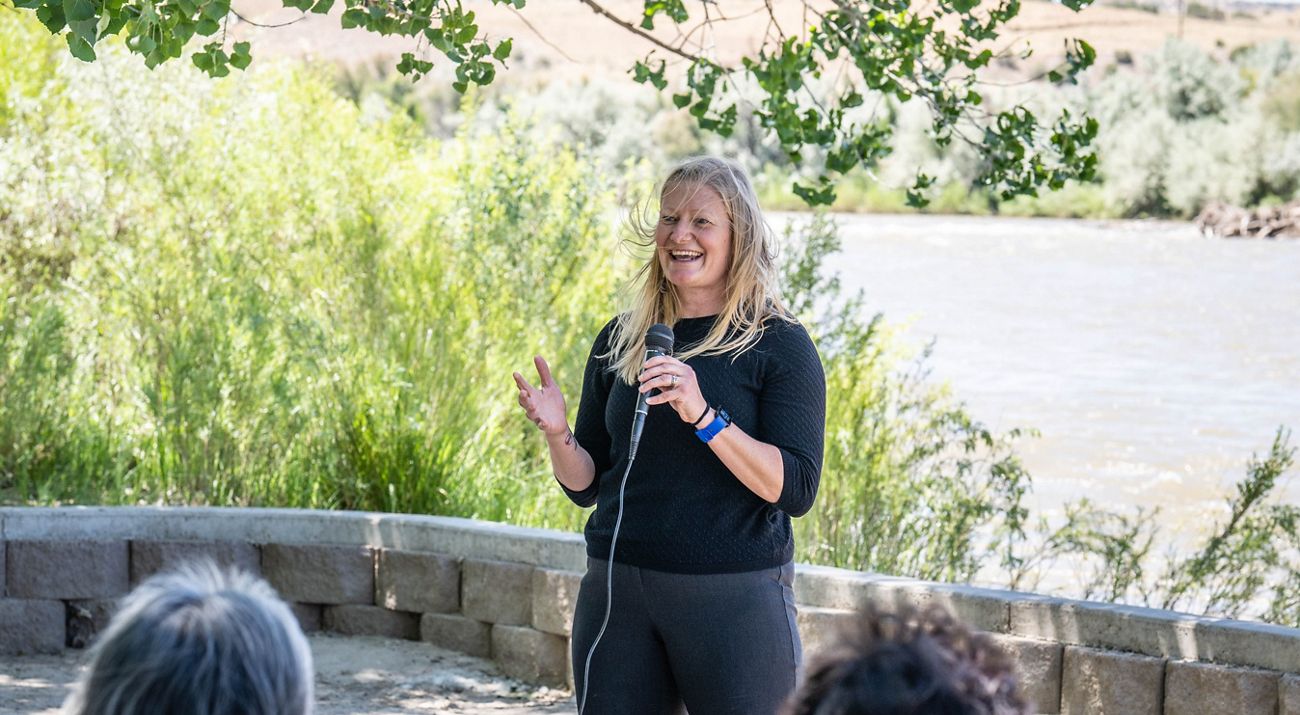 A blonde woman talking into a microphone in front of a group of people by a river.