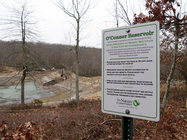 Sign alerting visitors about the demolition and closure of O'Conner dam. Large construction equipment is visible in the background opening  a breach in the center of the earthen dam.