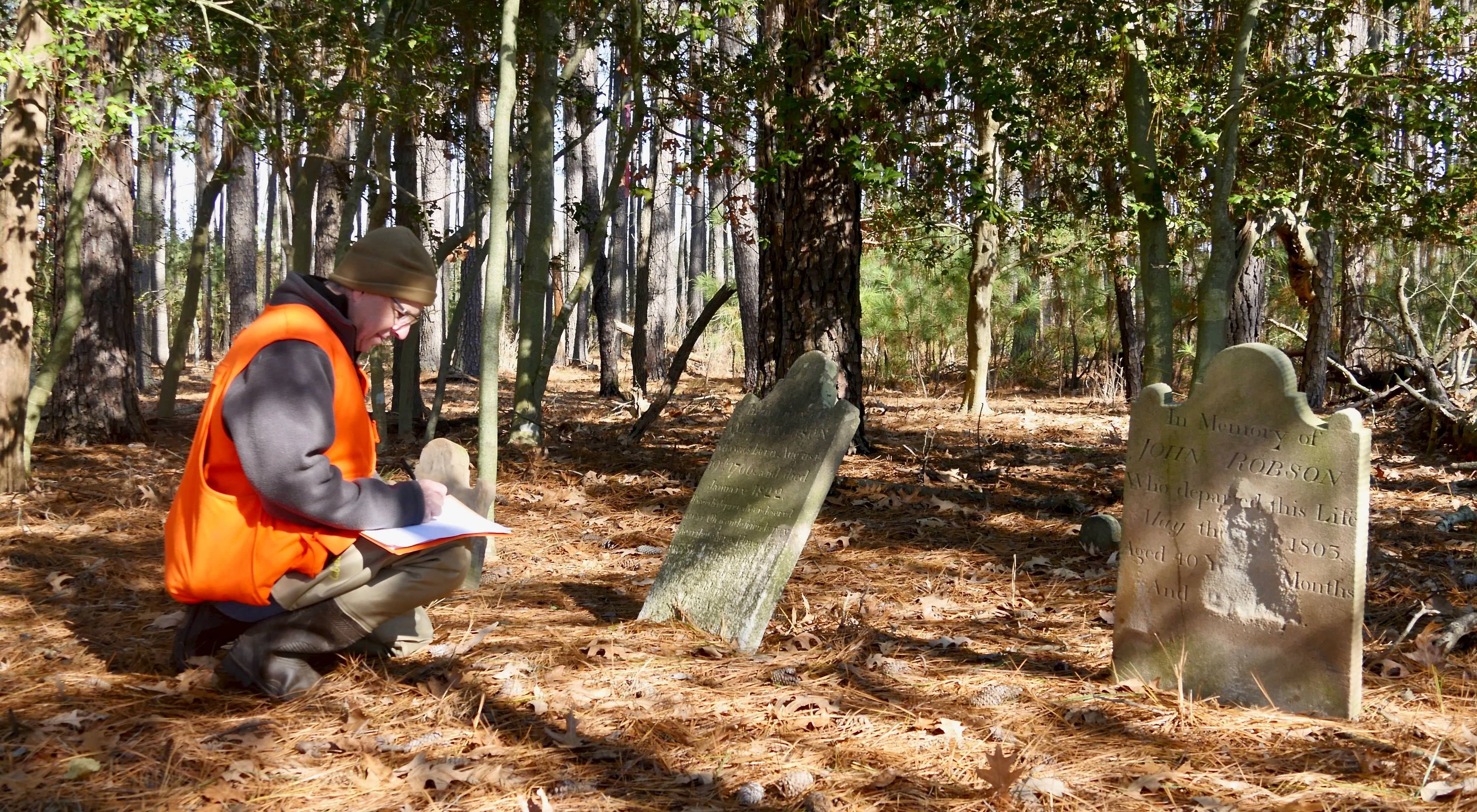 Man kneels with pen and paper in front of old tombstone in wooded area.