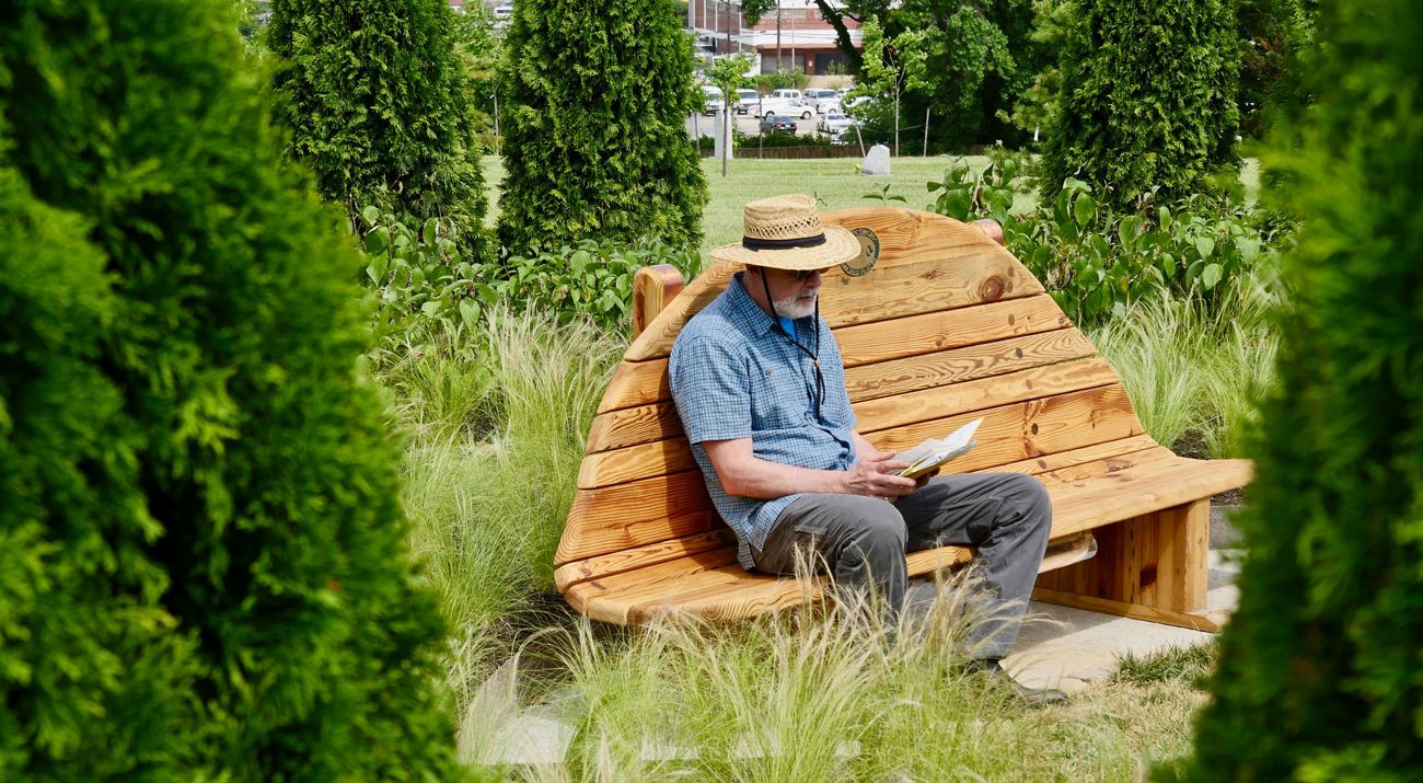 A man sits on a high-backed wooden bench reading a book in a green space encircled by trees.