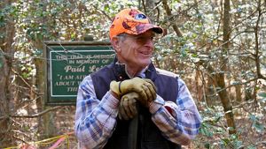 A smiling man rests his hands on the handle of a shovel.