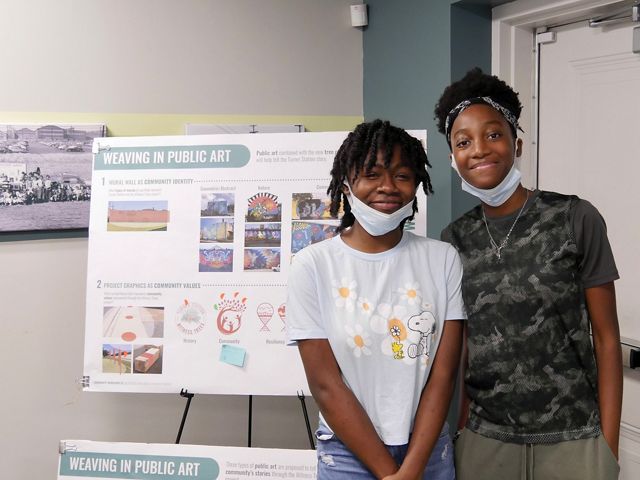 Two young people stand next to a display that reads weaving in public art during a community design workshop.