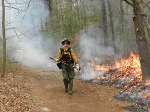 A photo of Natasha Whetzel walking on a path in the forest wearing yellow fire gear and holding an axe as a small flame burns to her right.