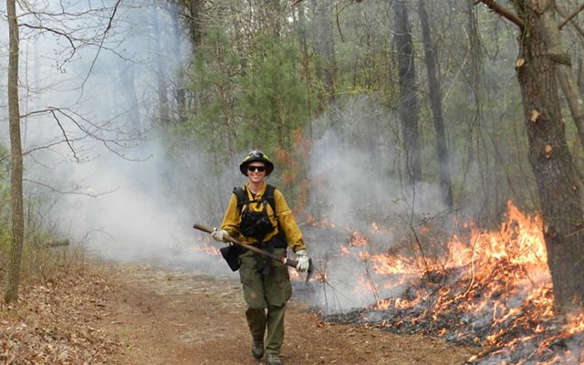 A person dressed in fire protective gear and holding an axe walks on a dirt path in a forest as a controlled fire burns behind them.