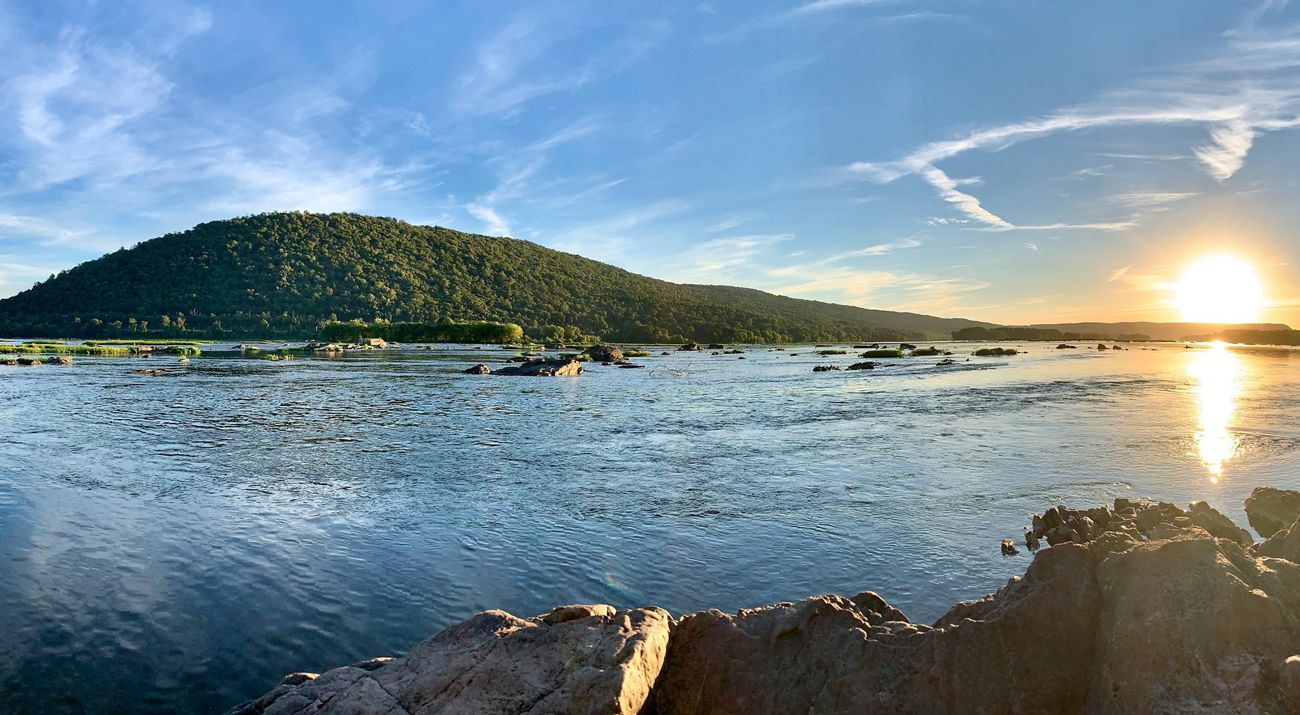 View of Hamer Woodlands at Cove Mountain from the east bank of the Susquehanna River. A large boulder is in the foreground. There are ripples on the surface of the water.