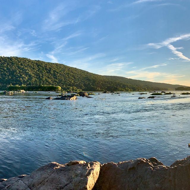 View of Hamer Woodlands at Cove Mountain from the east bank of the Susquehanna River. A large boulder is in the foreground. There are ripples on the surface of the water.