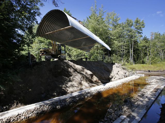 A large, corrugated metal half-pipe suspended over a ditch full of water.