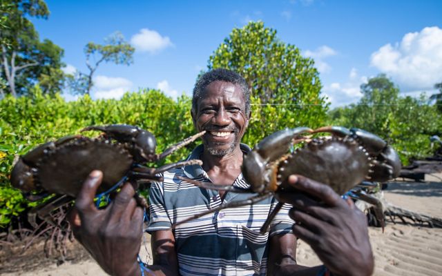 A crab fisherman smiles and holds up two crabs caught from a mangrove forest in Lamu County, Kenya.