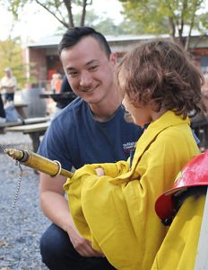Patrick Ma shows prescribed fire equipment to a child who wears yellow fire gear and holds a hose.
