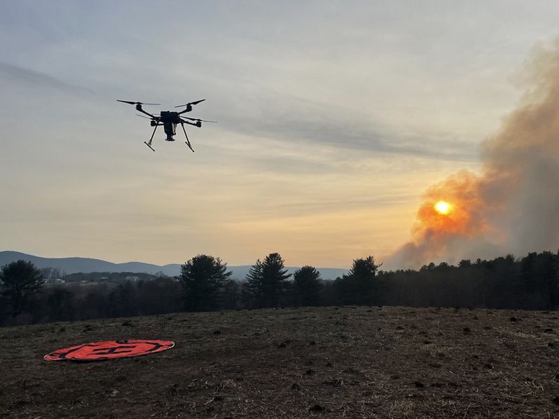 A drone flies above a field. Smoke is seen in the distance.