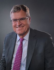 A man wearing a dark suit and purple tie smiles at the camera.