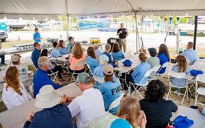 A group gathered seated under a tent, listening to a speaker, with oyster partnership banners visible.