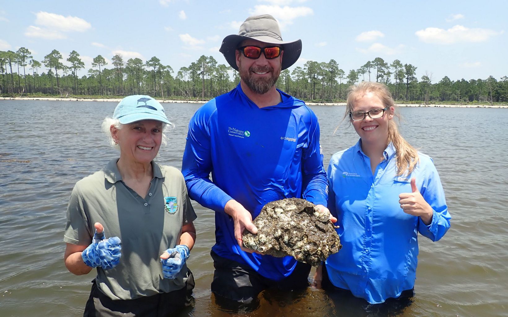 Three people stand smiling in waist-deep water holding rocks with new oysters grabbing hold.