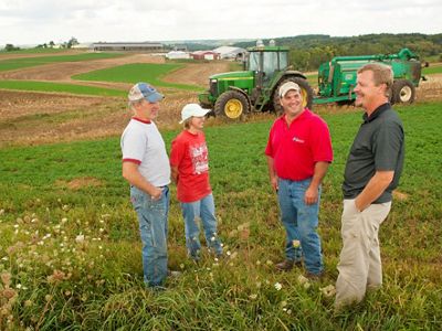 Four people standing in a farm field talking; a large green tractor is in the background.