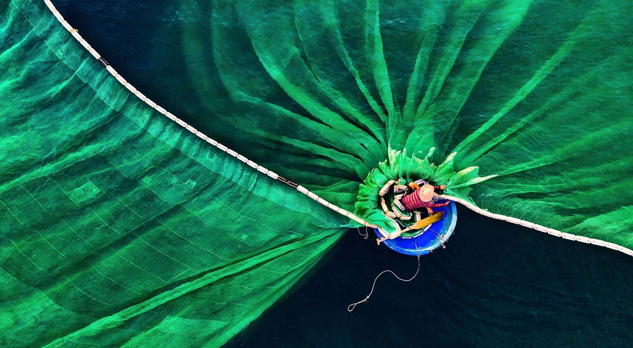 A person in a small circular boat casts a massive fishing net into the water.