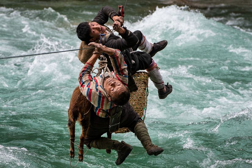 Two people use a rope and pulley system to cross a raging river with a basket and an animal.
