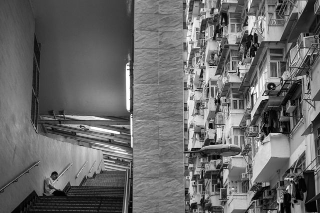 A black and white view of buildings in Hong Kong. On one side is a person sitting in a stairwell alone, and on the other side is dozens of balconies in a tall building.