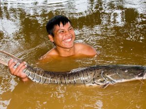 A Kichwa man holds a grey fish with both hands, inside a river.