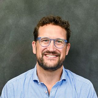 A headshot photo of a man wearing blue glasses and a blue checked shirt against a green-gray background.