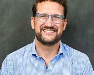 A headshot of a man wearing blue glasses and a blue checked shirt against a grey-green background.