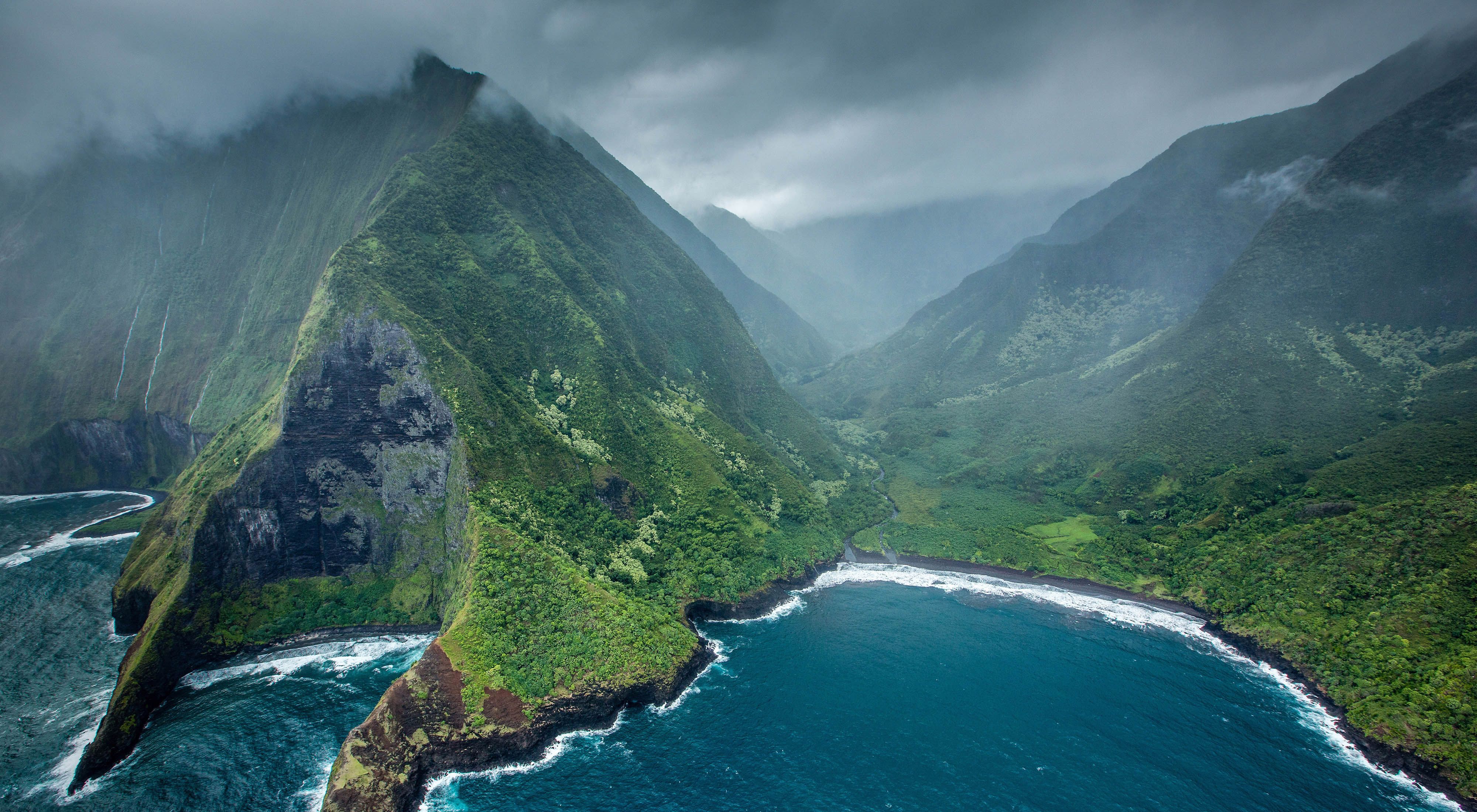 Aerial view of steep, craggy, green mountains jutting into blue ocean waters of Hawaii.