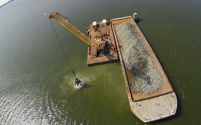 Aerial view looking down on a floating barge carrying crushed blocks of concrete. The grabber claw of a crane floating next to the barge dumps a load of concrete into the water.
