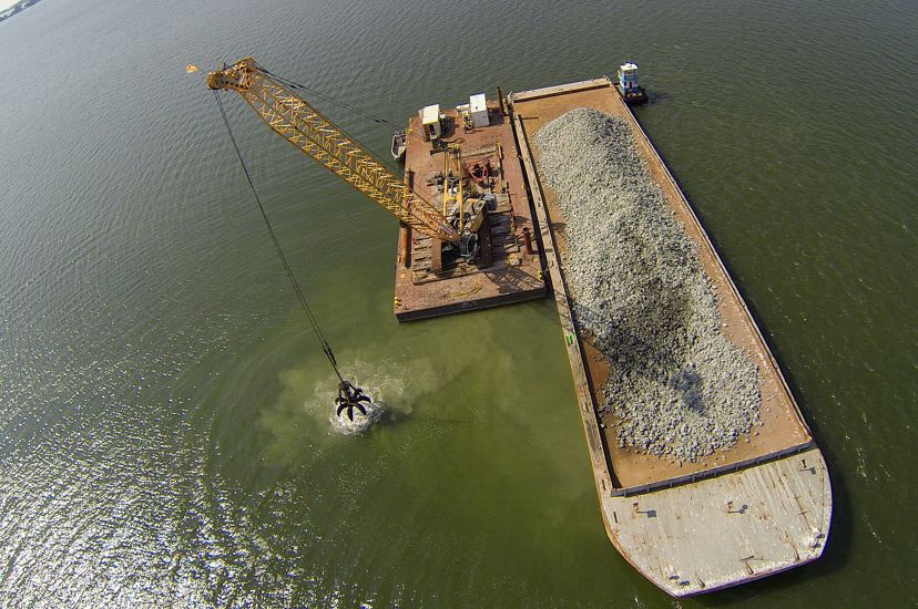 Aerial view looking down on a floating barge in the Piankatank River. A large crane is being used to scoop up chunks of granite rock from a pile on the barge and place them on a new oyster reef.