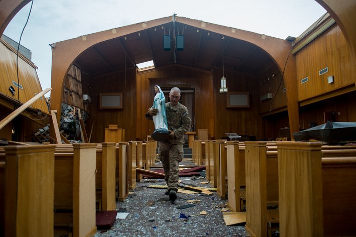 A man carries a large statue of the Virgin Mary. He walks down the center aisle of a small chapel that has had its roof torn off and is open to the sky. Splintered wood covers the floor.