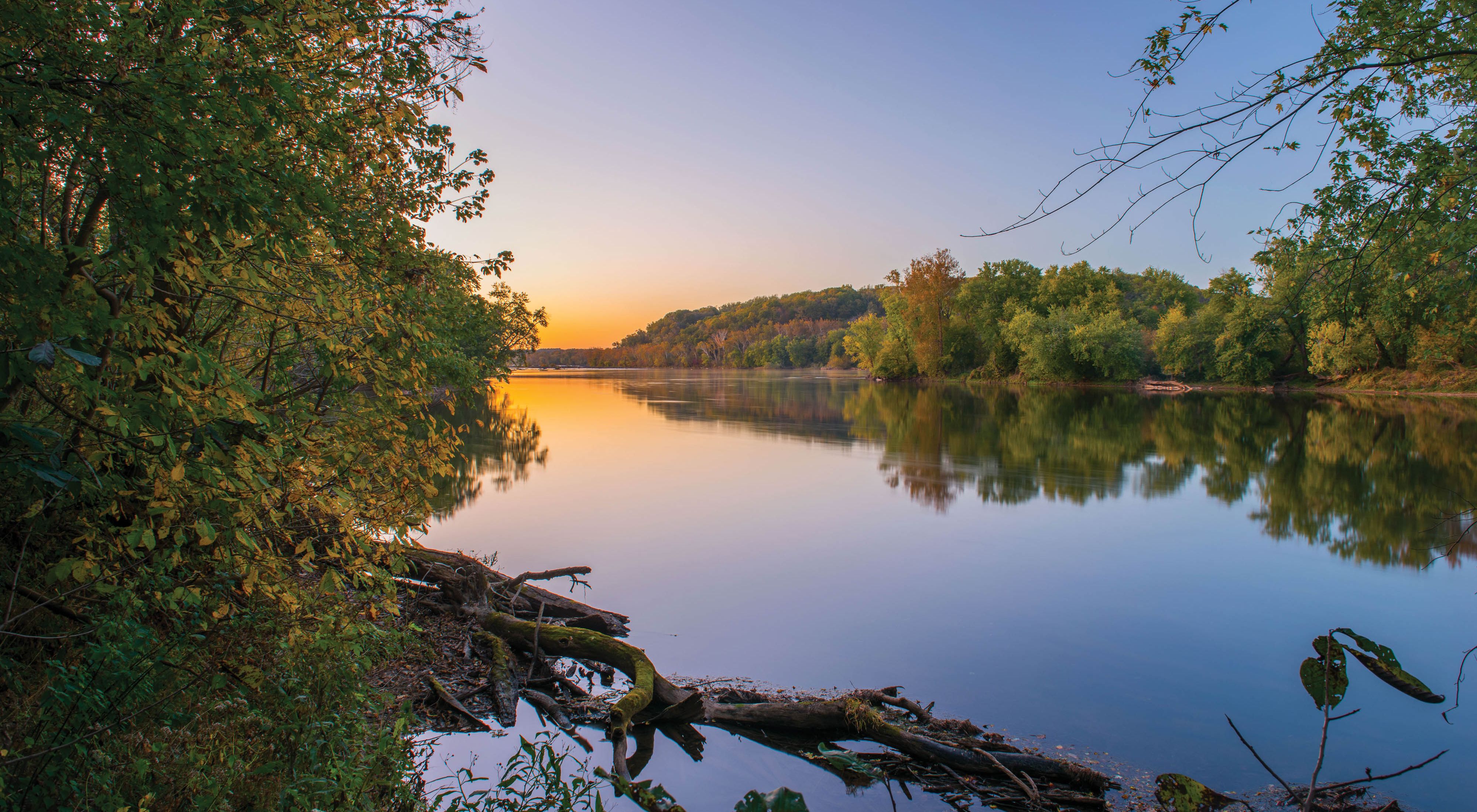 The Potomac River runs through Virginia's Fraser Preserve. The tree lined banks are reflected in the mirror-like surface of the water as the horizon glows in the light of the rising sun.