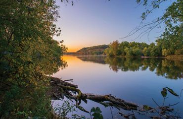 The sun rises over the flat, glassy surface of the Potomac River as it flows through TNC's Fraser Preserve. The wide river channel is lined with tall mature trees.