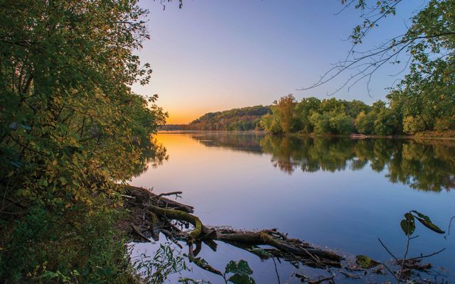 The sun rises over the flat, glassy surface of the Potomac River as it flows through TNC's Fraser Preserve. The wide river channel is lined with tall mature trees.