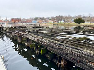Broken timbers stretch across a narrow urban waterway with parkland and historic buildings in the background.