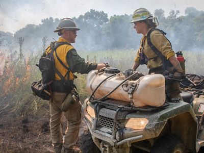 A man and women in fire clothing stand near an ATV while a prescribed fire burns the field behind them.