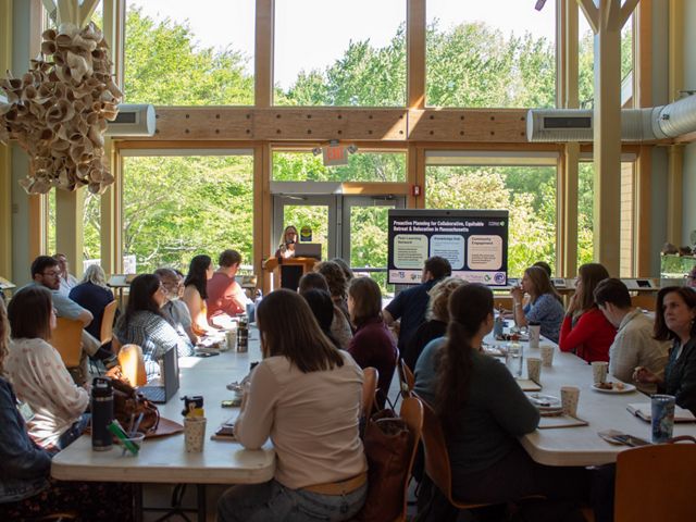 Workshop attendees sitting at long tables. A presenter stands at the front of the room next to a screen displaying a powerpoint.