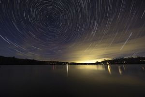 Time lapse photo of stars circling in the night sky over the calm surface of a small pond