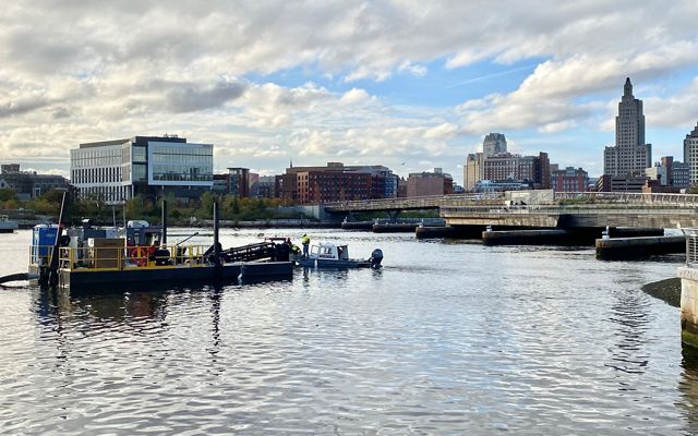 A dredge barge sits in the Providence River with a graceful pedestrian bridge and the city's skyline in the background.