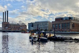 A dredge barge floats on the water in low light with the Providence skyline in the background