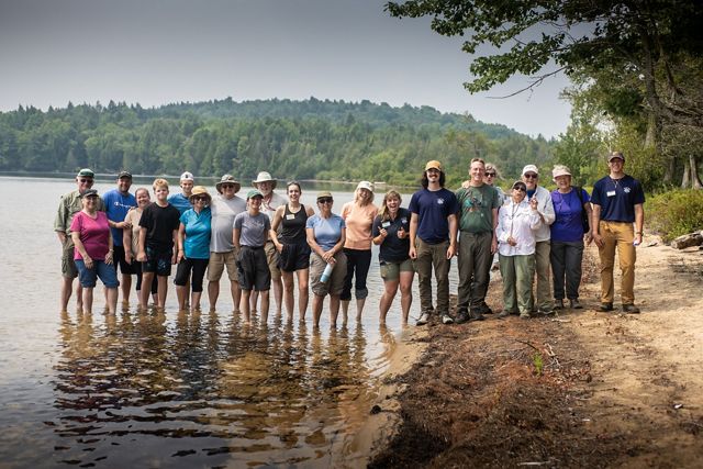 21 people stand on the sandy shore and in the water at Follensby Pond.